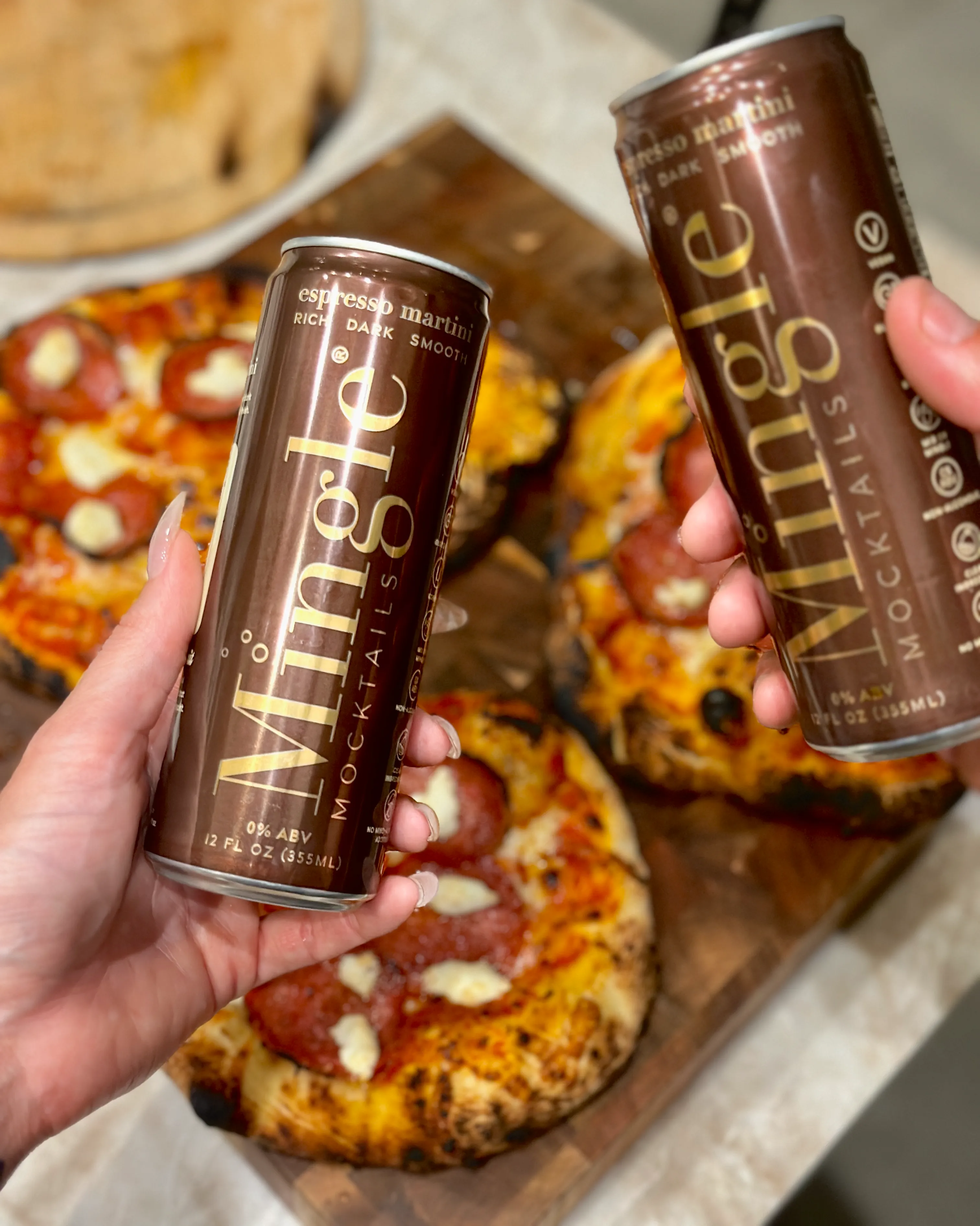 Two hands hold cans of Mingle Mocktails Espresso Martini in front of small pepperoni pizzas on a board.