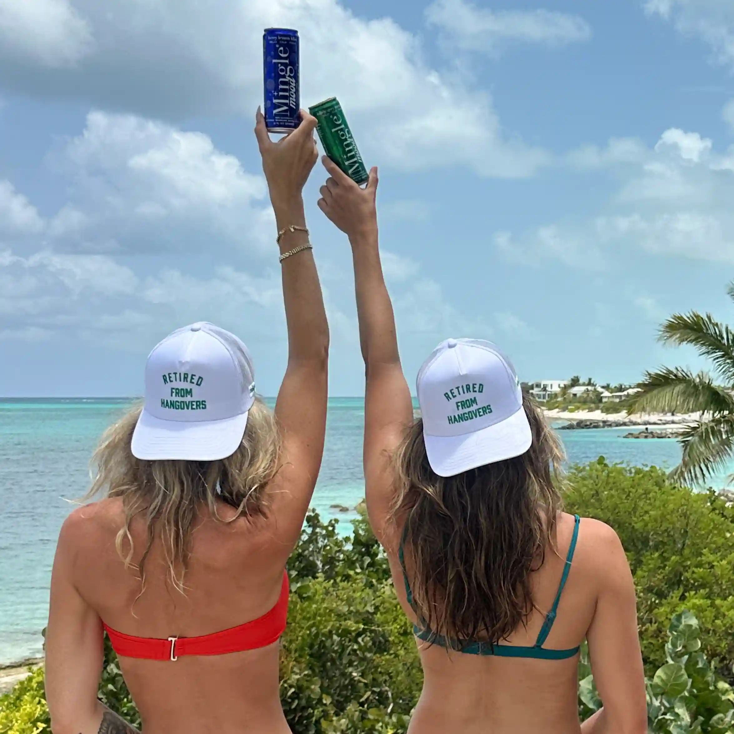 Two women wearing 'Retired from Hangovers' hats hold up beverage cans overlooking a tropical ocean.