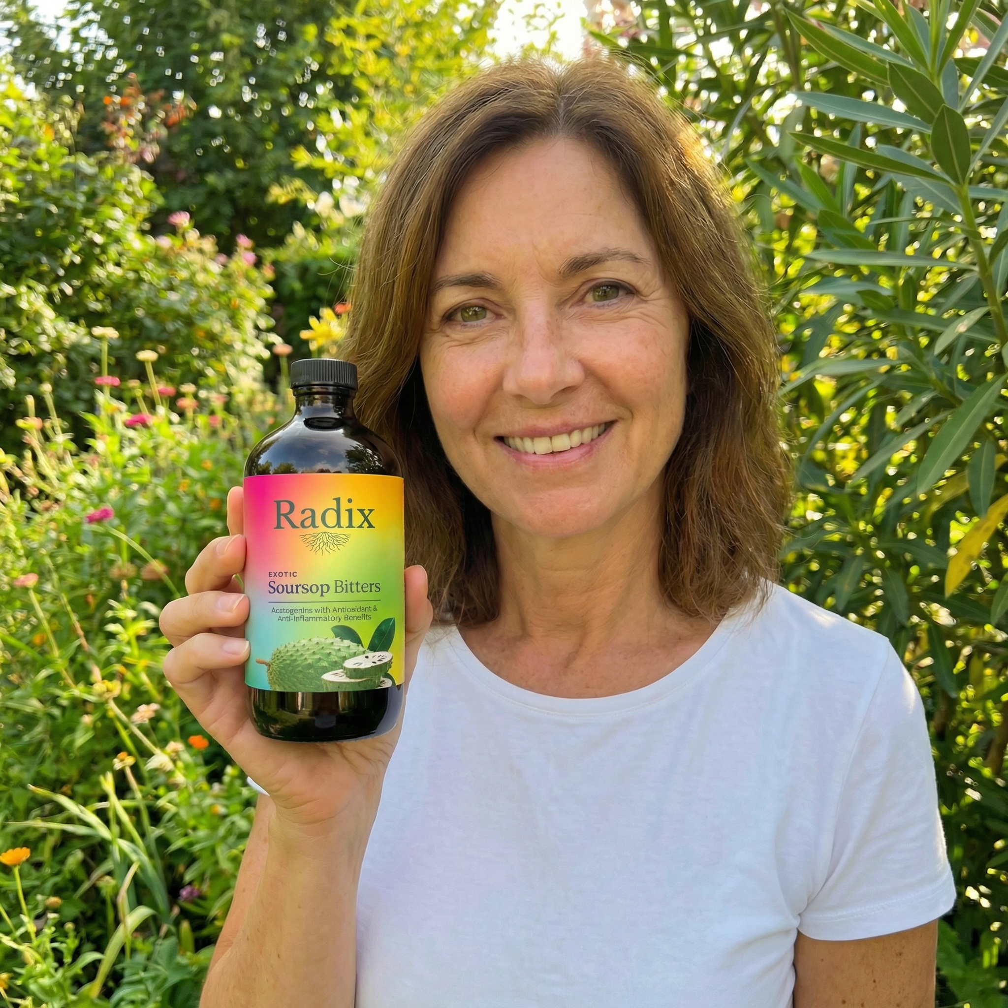 A smiling woman in a white t-shirt holds a bottle of Radix Soursop Bitters in a garden.