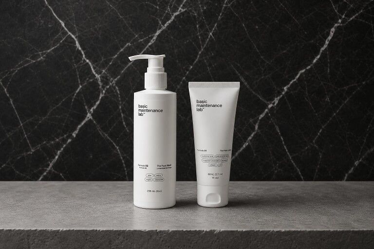 Two white cosmetic containers from 'basic maintenance lab' on a stone surface against a black marble background.