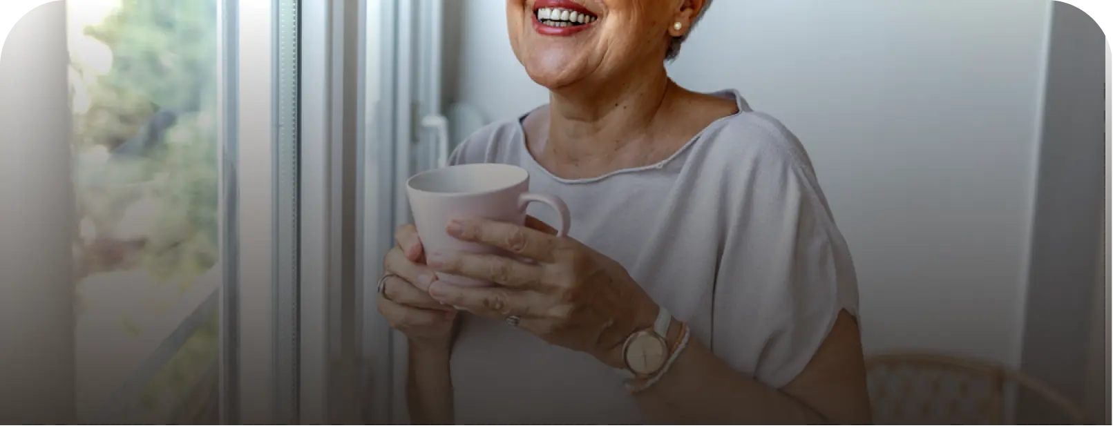 A smiling older woman in a white shirt holds a white mug by a window.