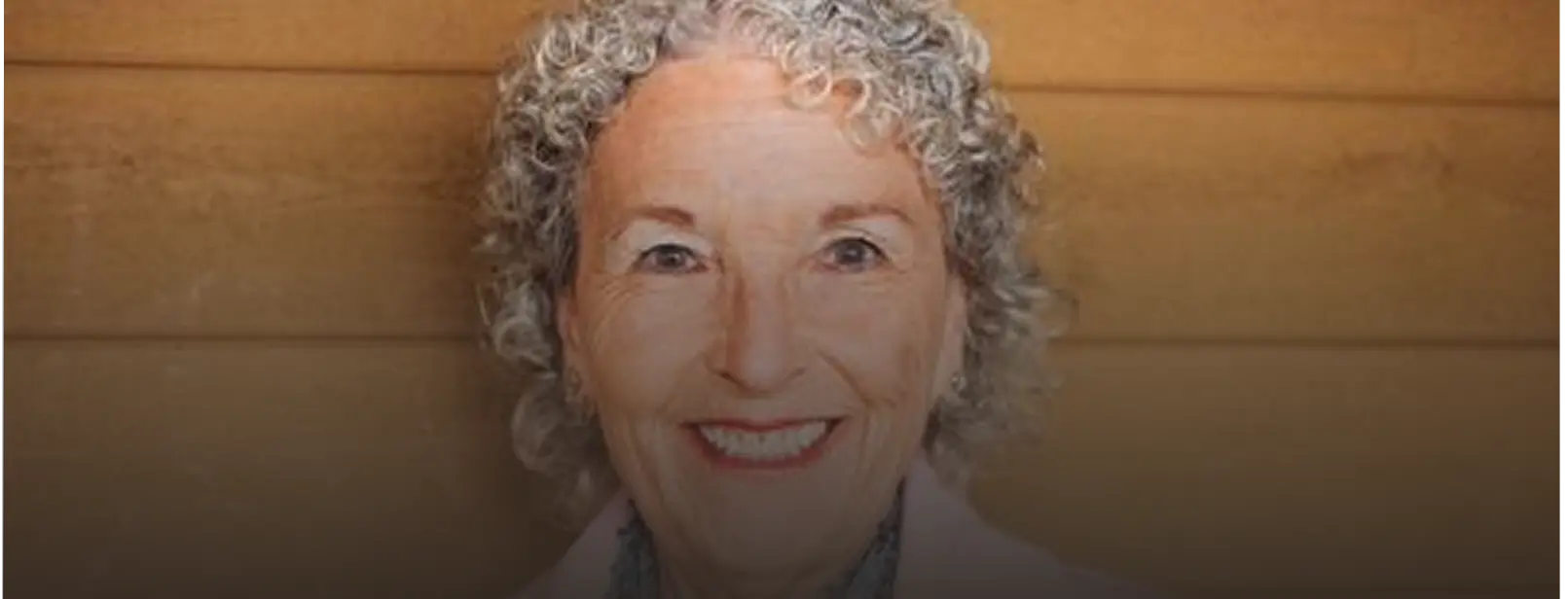 A smiling older woman with curly gray hair looks at the camera against a wooden background.