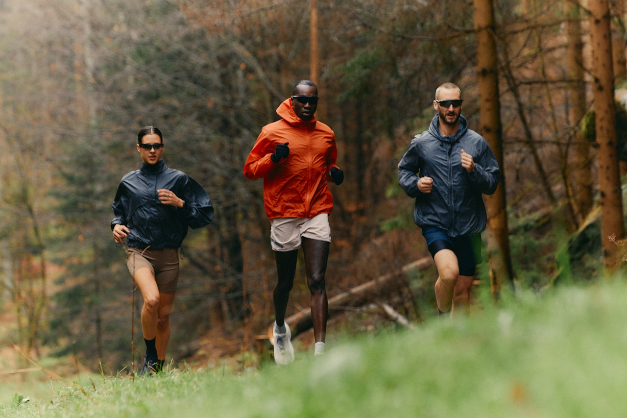 Three people jogging in a forest wearing jackets and sunglasses.