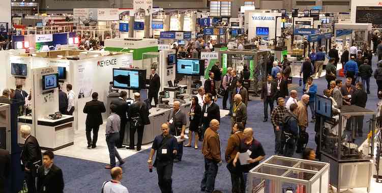 A busy convention hall floor filled with people walking among various trade show booths and exhibits.