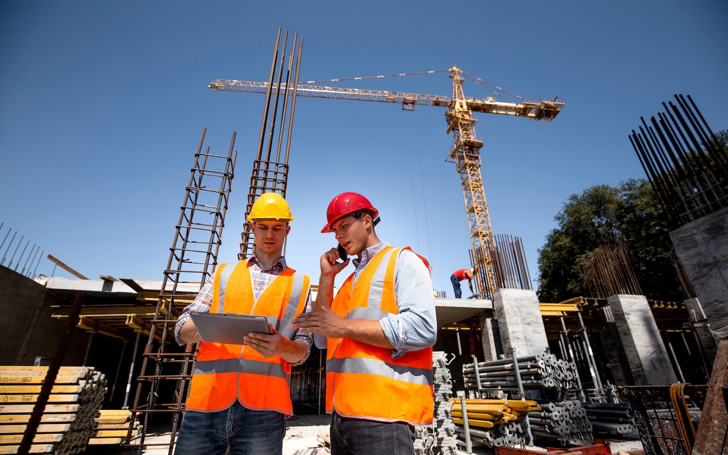 Two construction workers in safety gear consult a tablet and phone on a busy construction site with a crane.