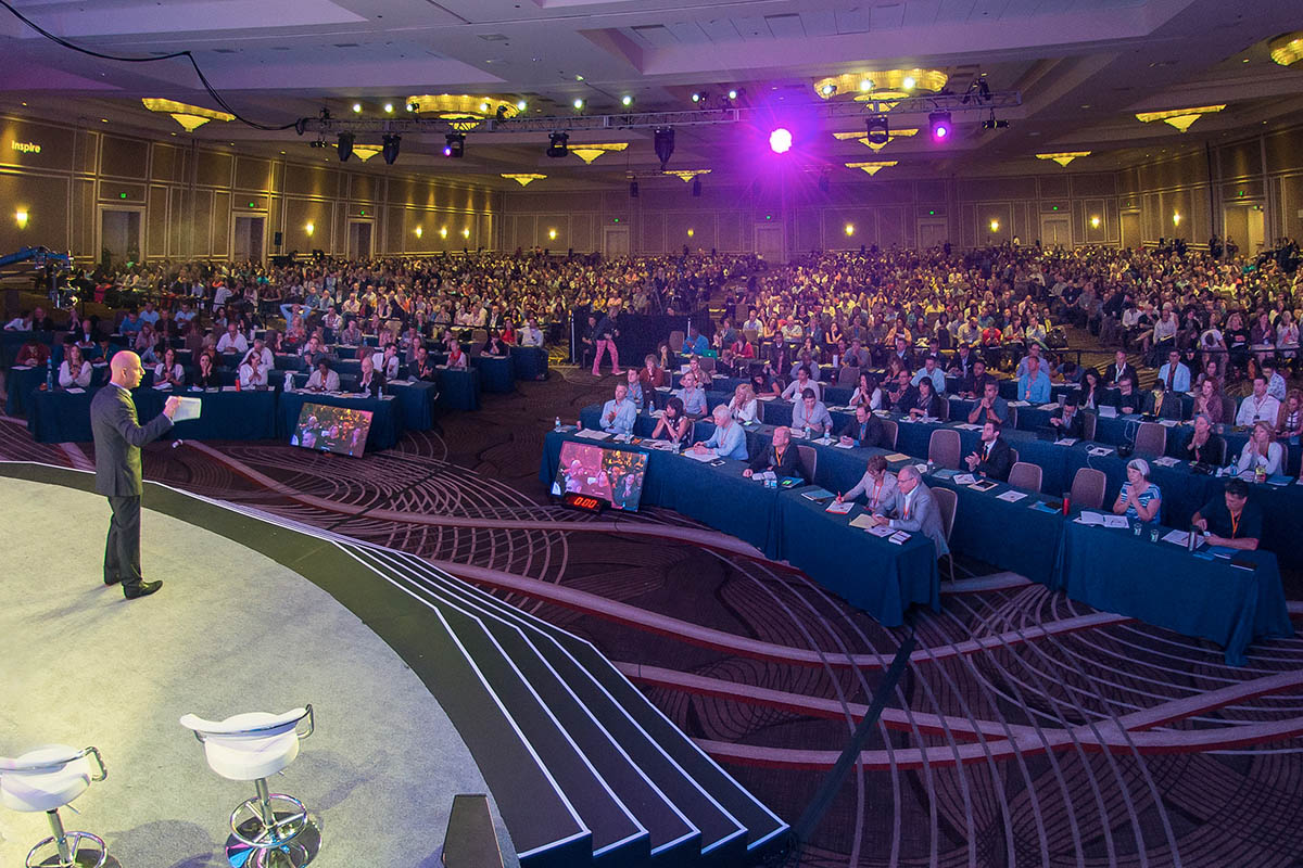 A speaker on stage addresses a large audience seated at tables in a conference hall.