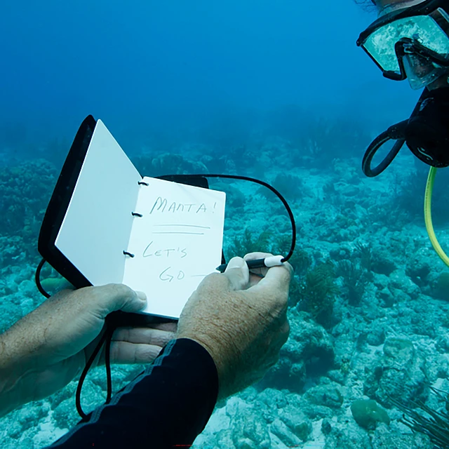 Diver holding notebook underwater with notes; another diver visible.