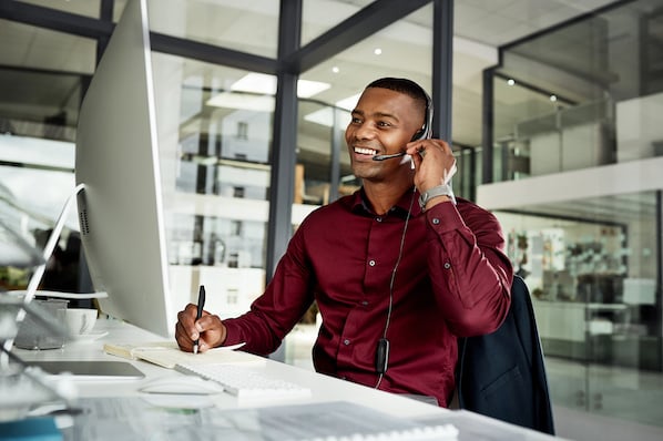 A smiling man with a headset sits at an office desk, looking at a computer and writing.