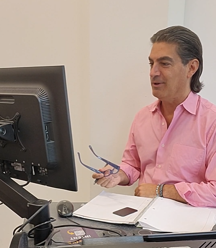 A man in a pink shirt sits at a desk, holding glasses and looking at a computer monitor.