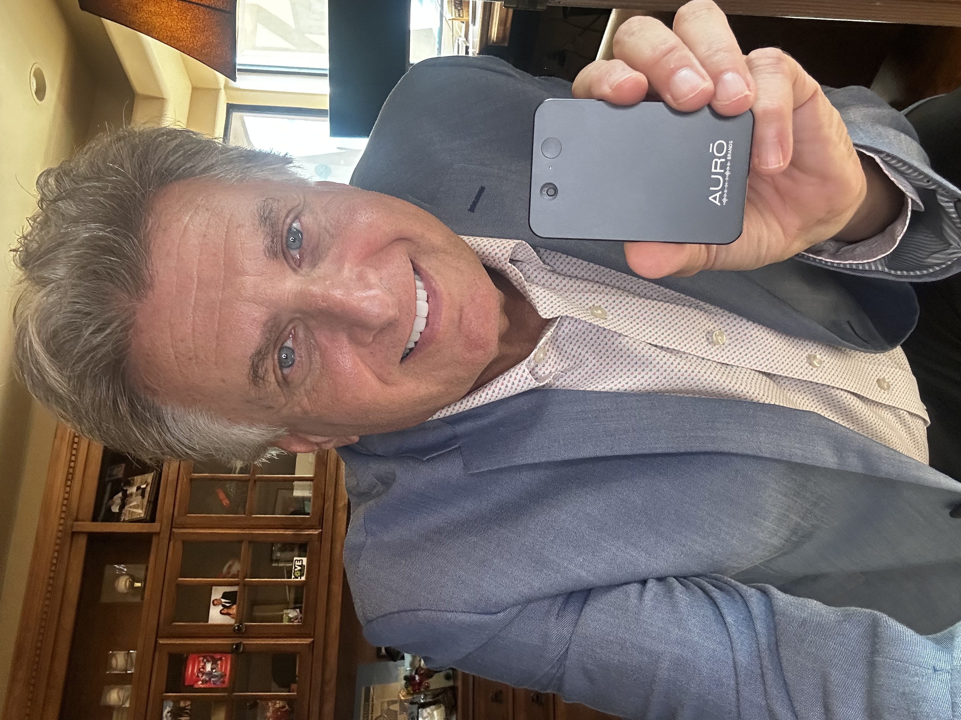 A smiling man in a light blue blazer holds up a small black electronic device.