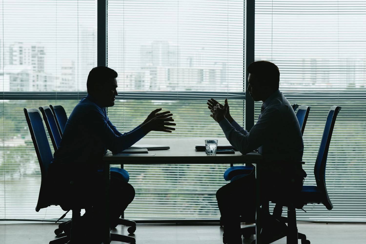 The silhouettes of two men sitting at a table and talking in front of a large window.