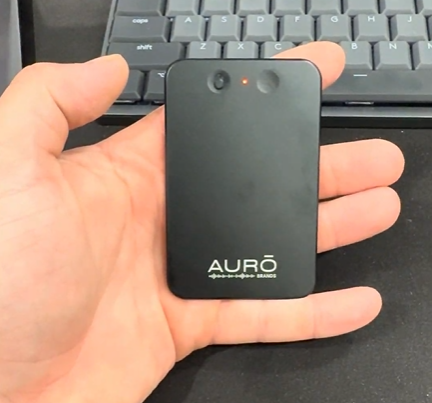A hand holding a small black electronic device with the Aurō logo in front of a keyboard.