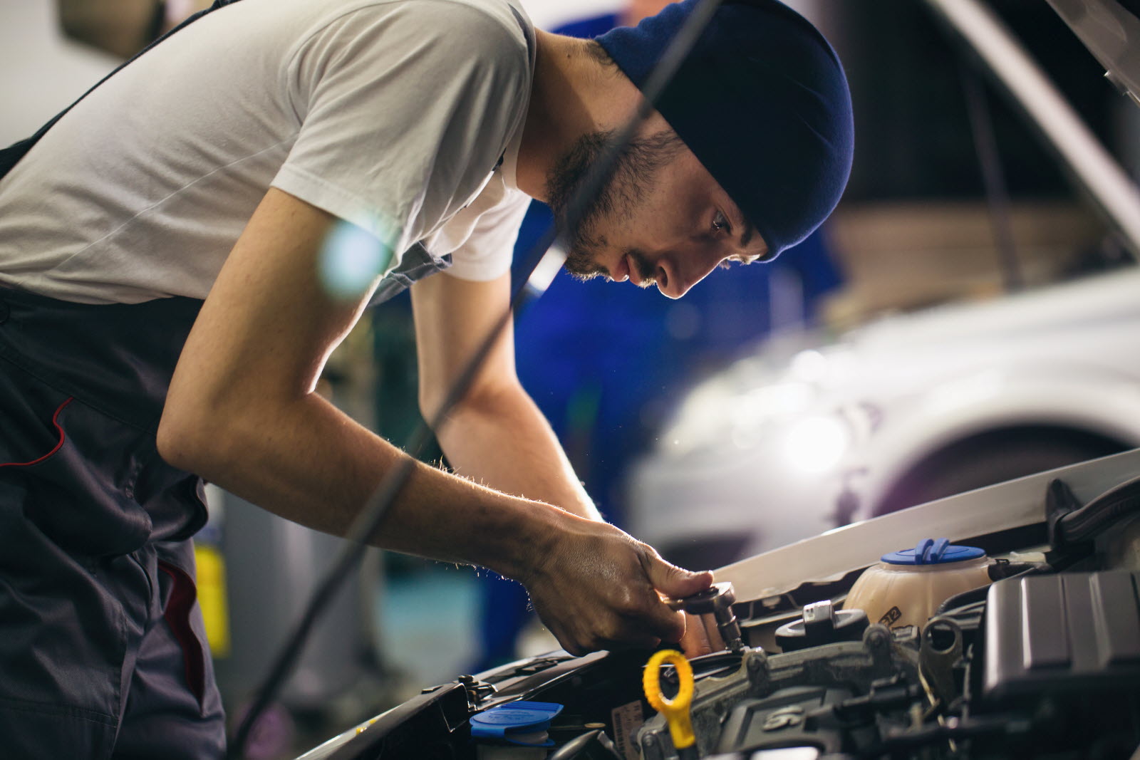 A mechanic wearing a beanie leans over an open car hood, working on the engine with a tool.