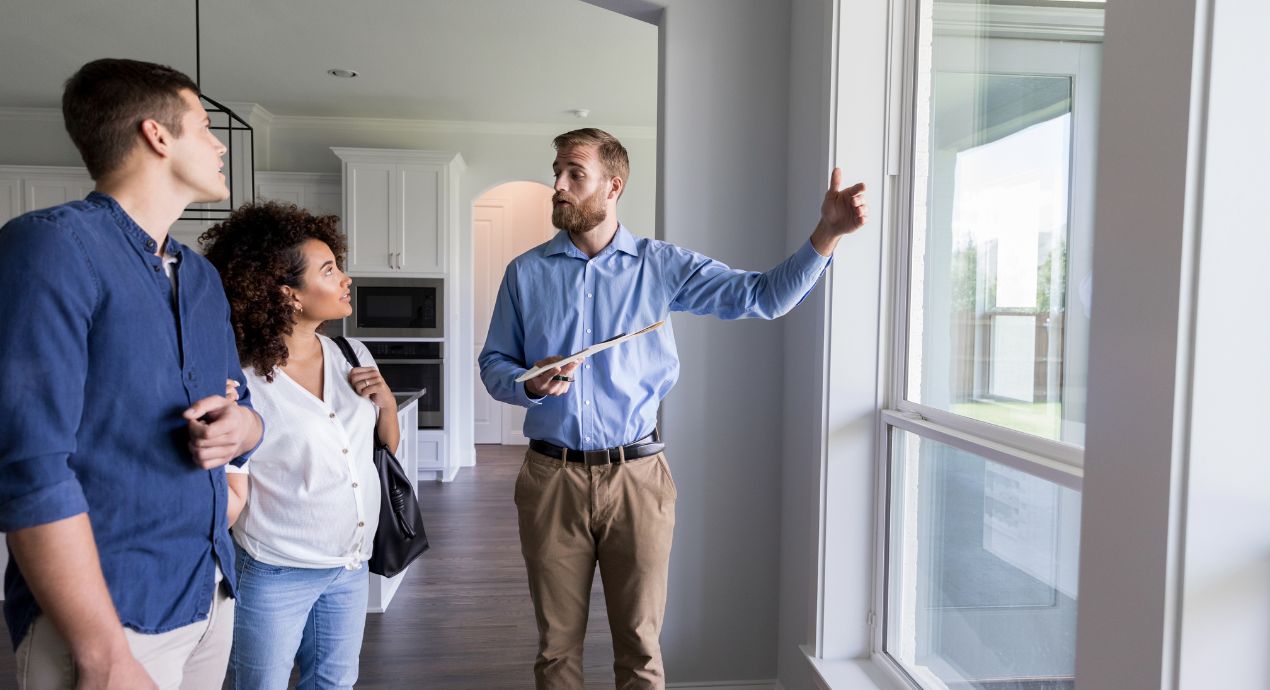 A real estate agent gestures toward a window while showing a house to a young couple.