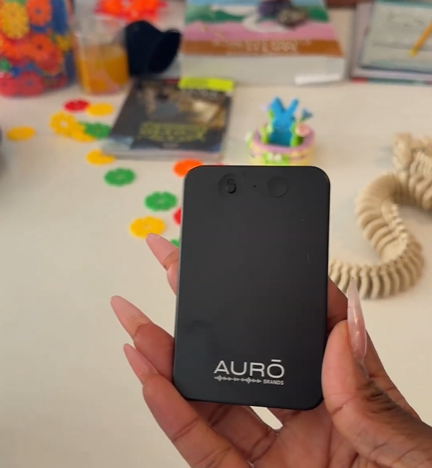 A hand with long nails holds a small, black rectangular device from Aurō Brands over a desk.
