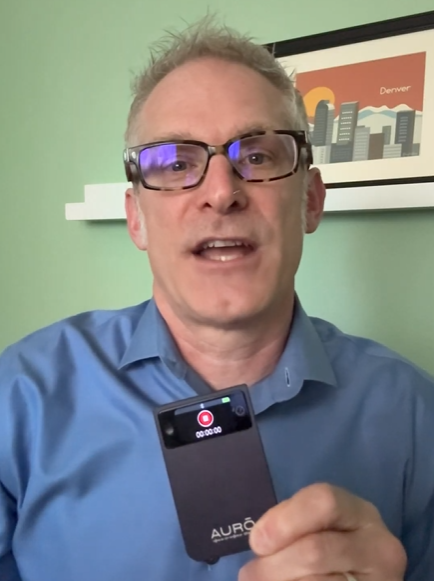 A man in glasses and a blue shirt holds a small black recording device in front of him.