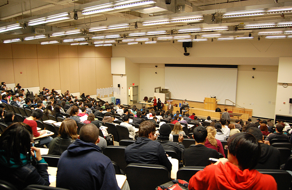 A large lecture hall filled with students facing a stage with a projector screen and speakers.