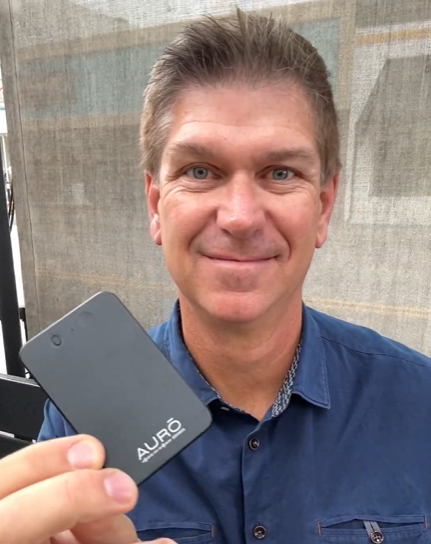 A smiling man in a blue shirt holds up a small, black rectangular device with the word AURŌ.