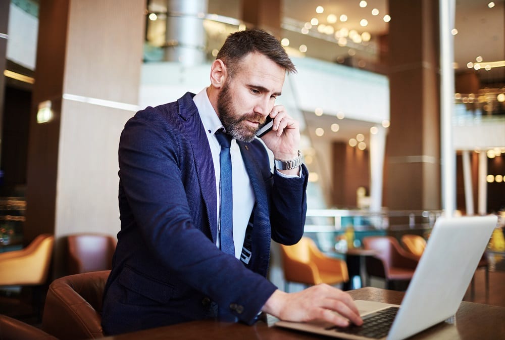 A man in a blue suit and tie talks on his phone while using a laptop computer.