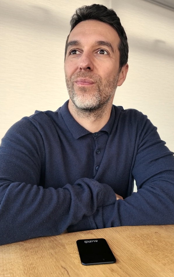 A man in a blue polo shirt sits at a wooden table, looking up and to the side.