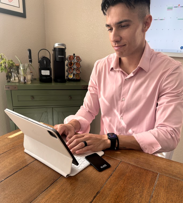 A man in a pink shirt sits at a wooden table, typing on a white tablet computer.