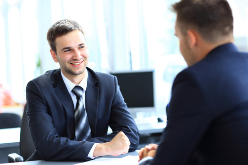 Two men in business suits sit across from each other at a desk in an office setting.