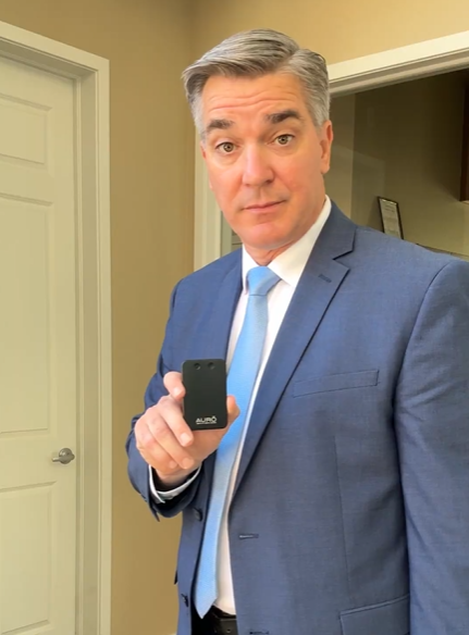A man in a blue suit and light blue tie holds up a small, black electronic device.