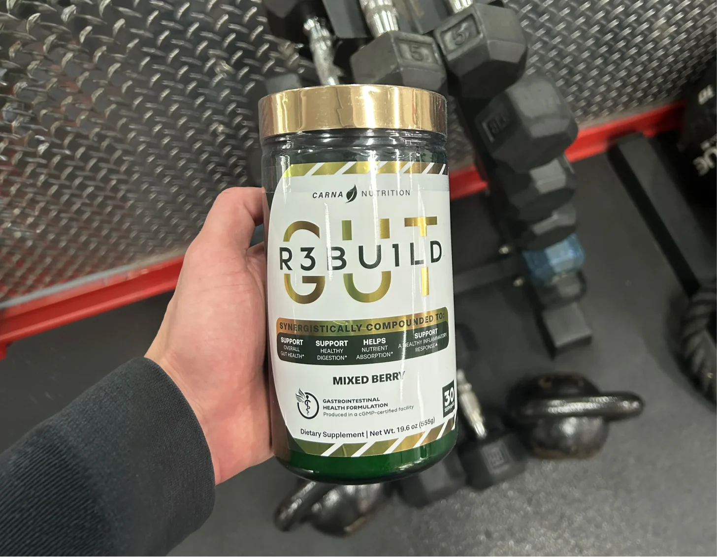A hand holding a jar of Carna Nutrition Gut R3build dietary supplement in front of a dumbbell rack.