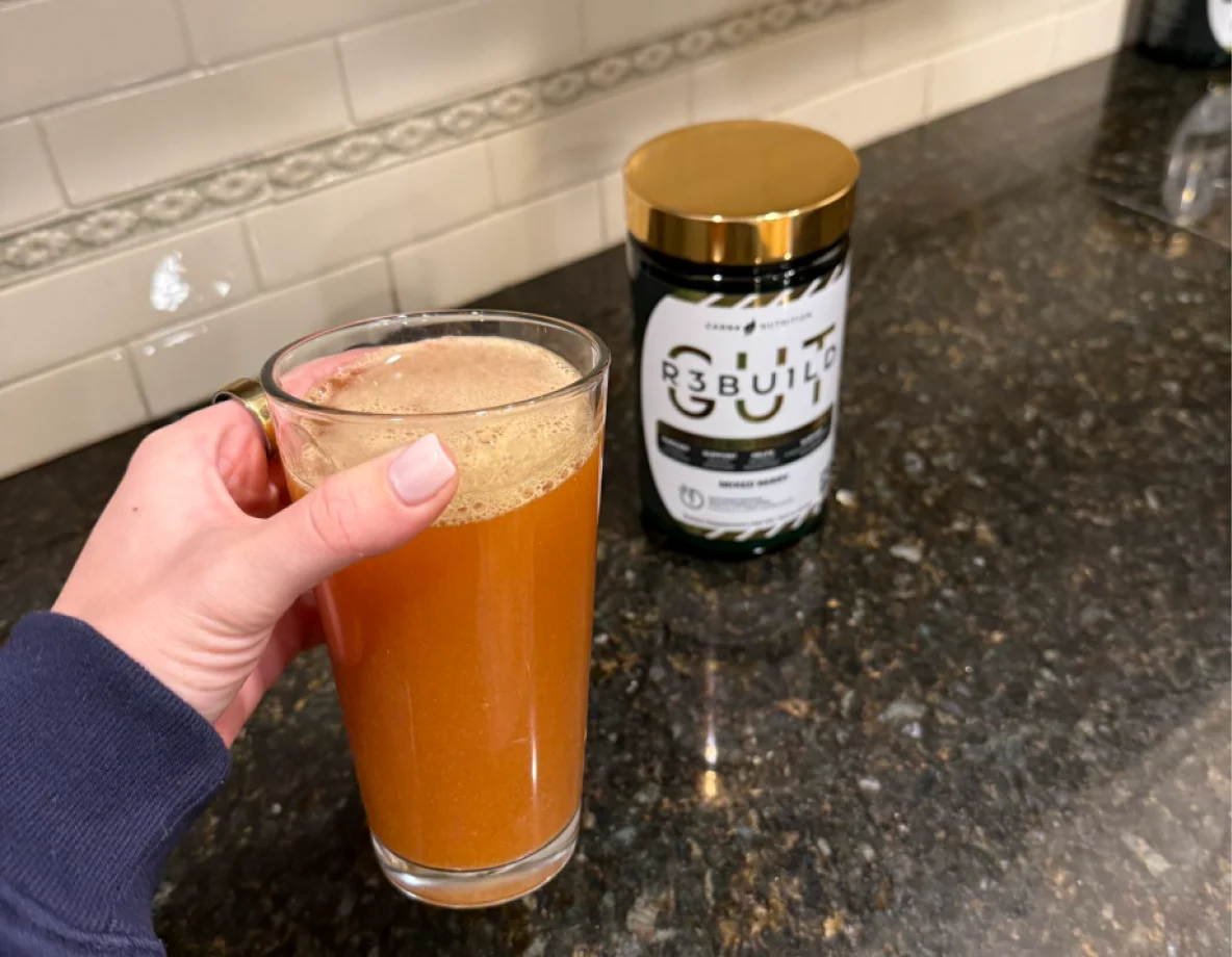 A hand holds a glass mug with a frothy orange beverage next to a supplement container.