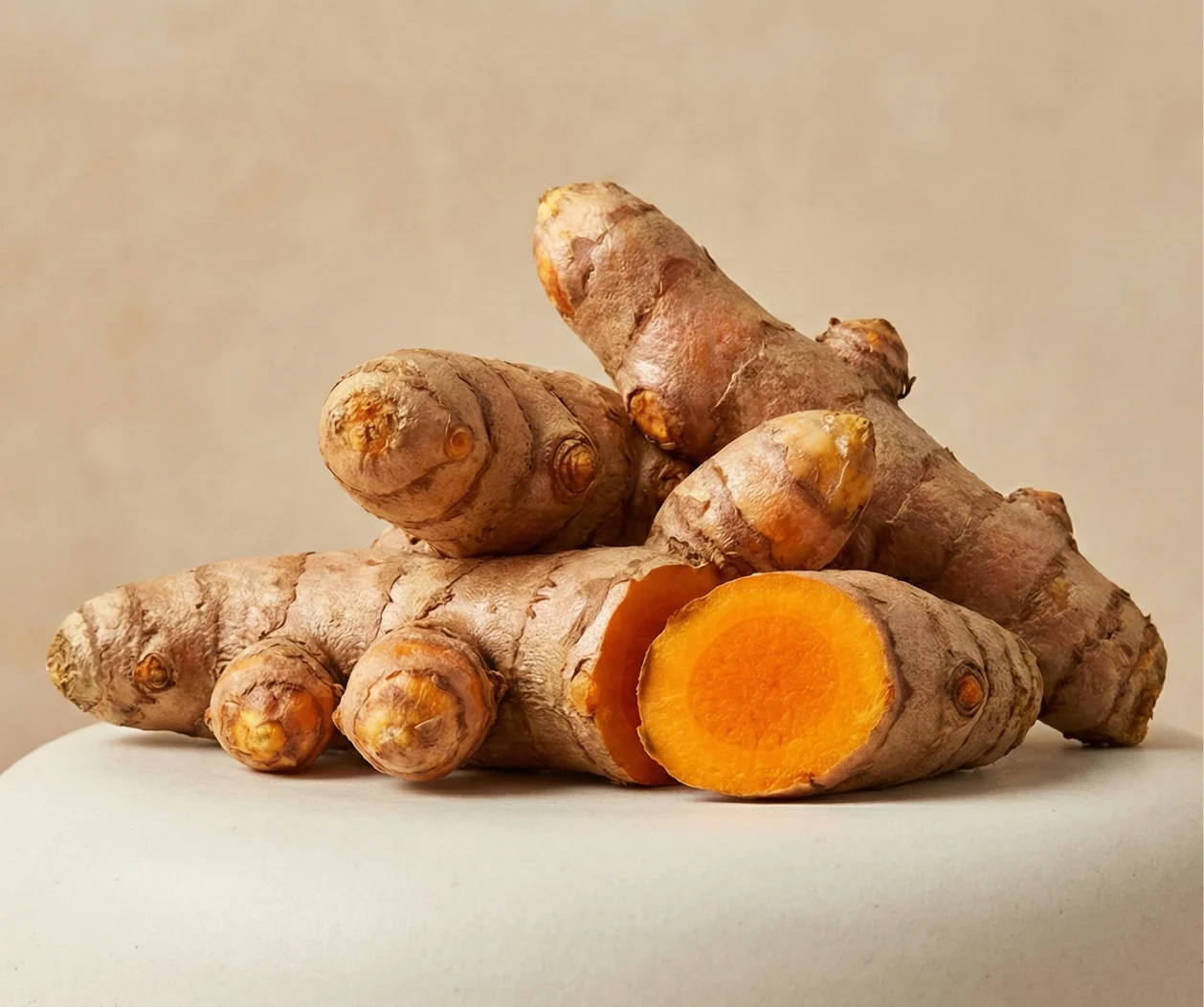 A pile of whole and sliced raw turmeric root sits on a light-colored surface against a tan background.