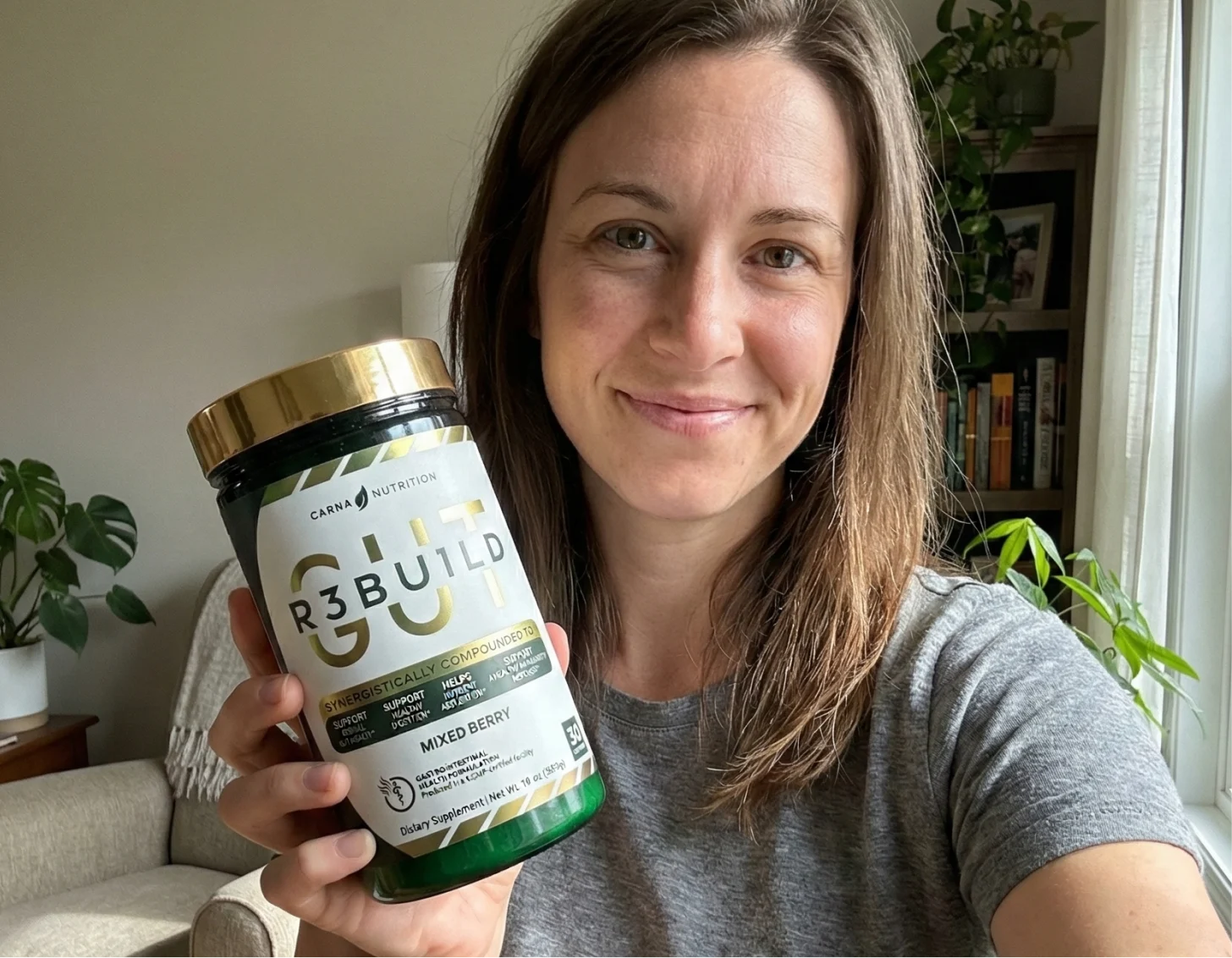 A smiling woman in a grey t-shirt holds up a container of Carna Nutrition Gut R3build supplement.