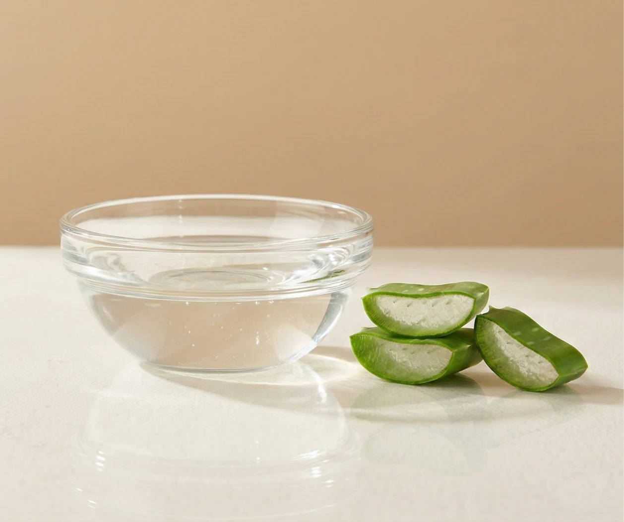 A small glass bowl of clear liquid sits next to three cut pieces of an aloe vera leaf.