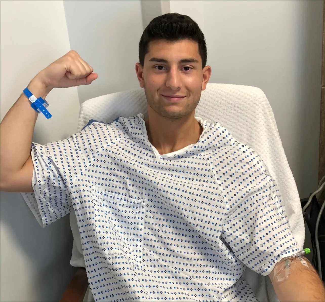 A young man in a hospital gown smiles and flexes his arm while sitting in a chair.
