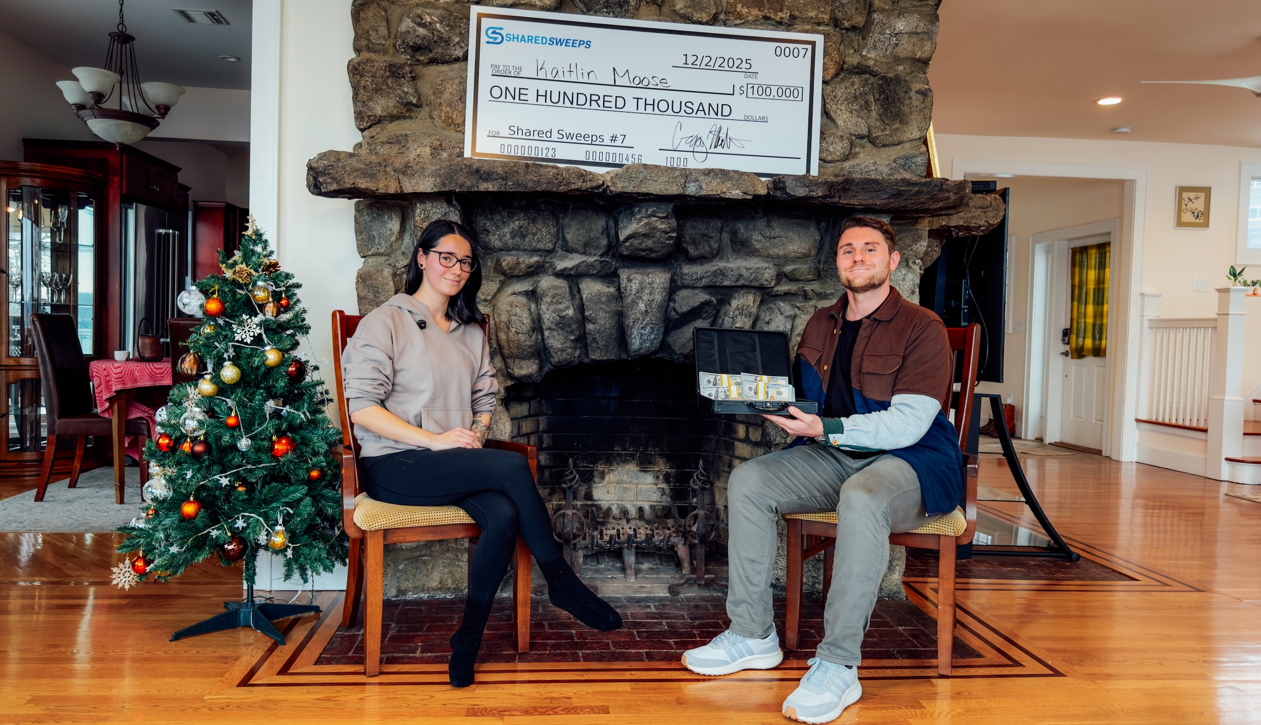 Two people sitting by a fireplace with a large check and a Christmas tree nearby.