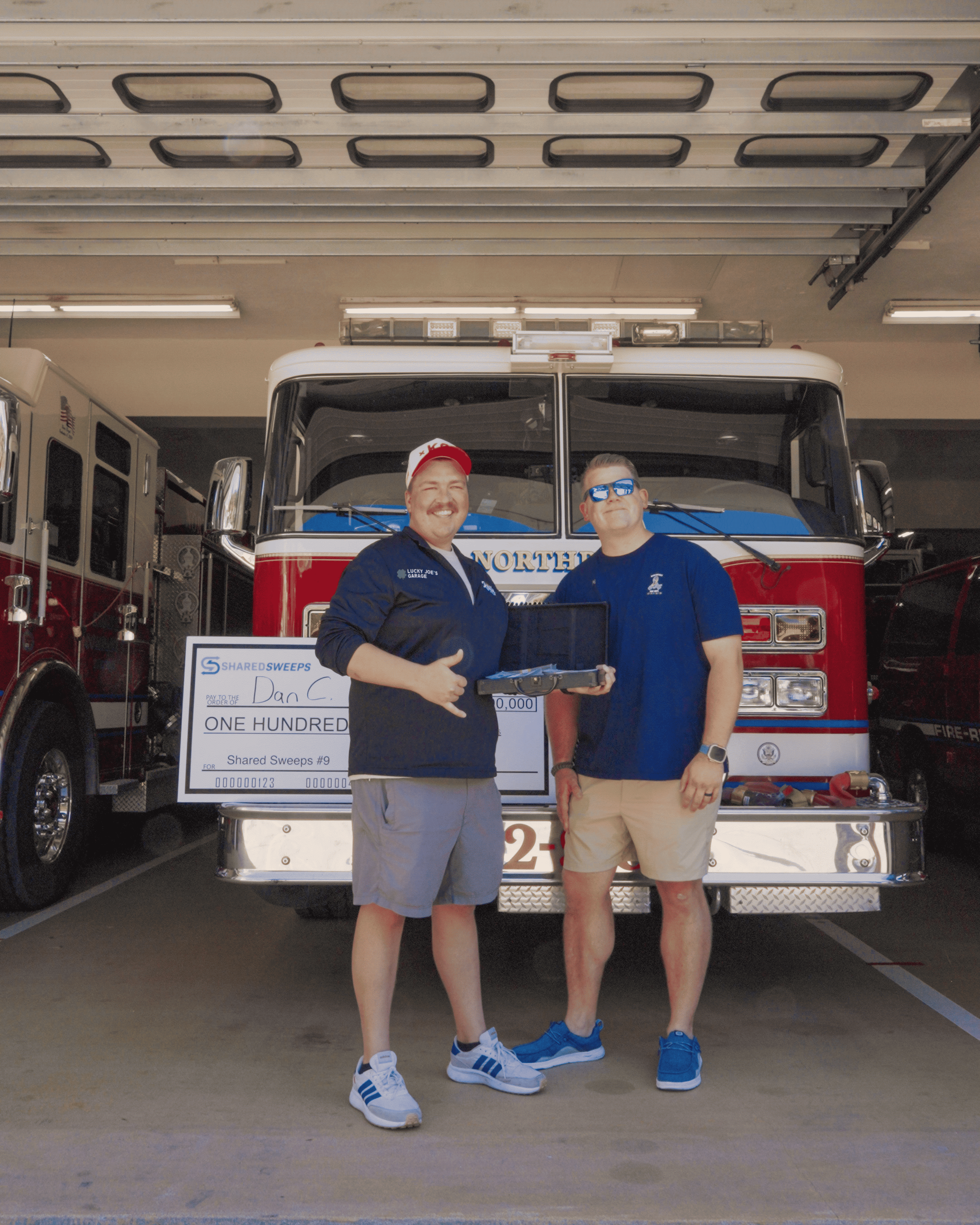 Two men pose with a large check and a briefcase full of cash in front of a fire truck.