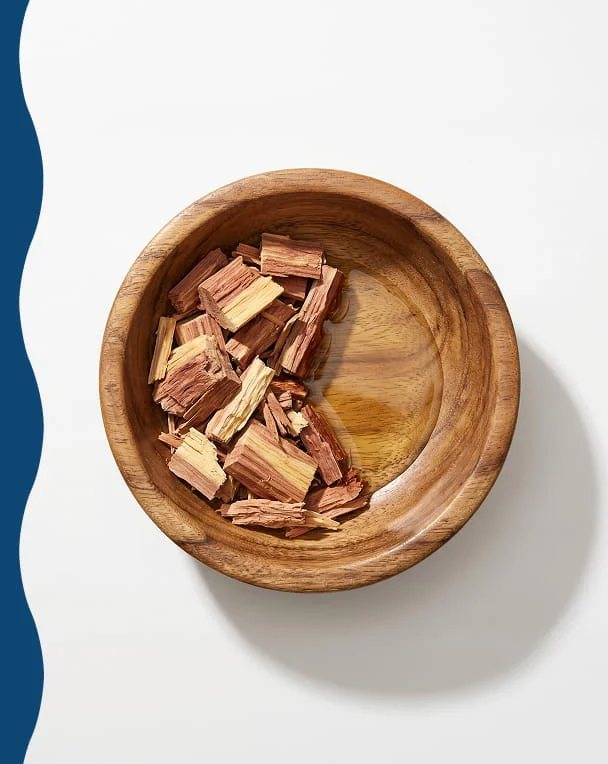 An overhead view of a wooden bowl filled with reddish wood chips on a white surface.