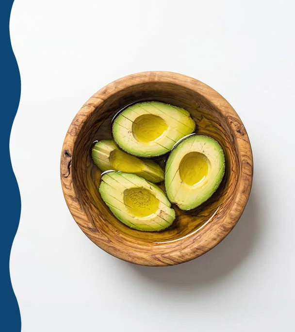 An overhead shot of several pitted avocado halves floating in oil inside a wooden bowl.