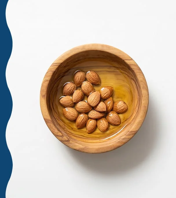 An overhead view of almonds soaking in a clear liquid within a light-colored wooden bowl on a white surface.