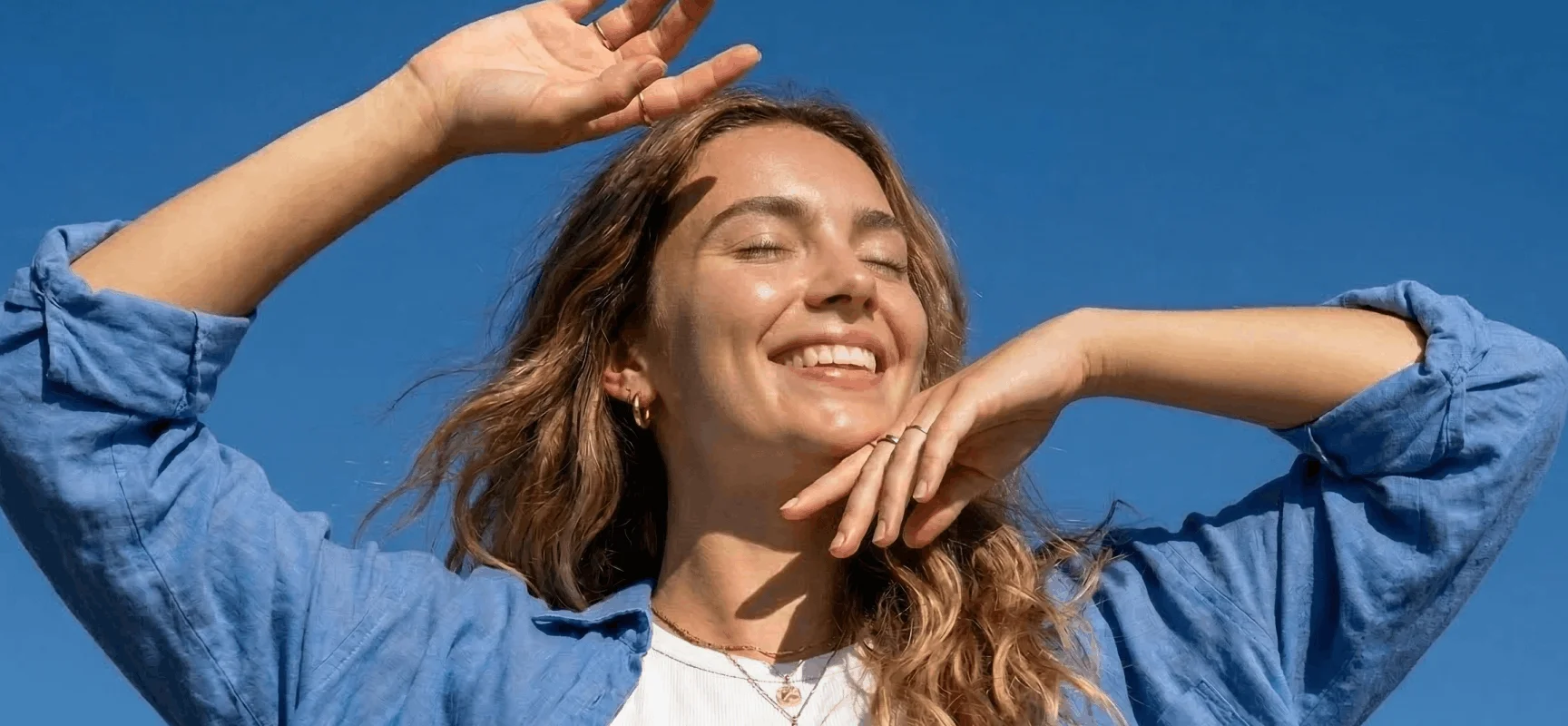 A woman with her eyes closed, smiling with her hands up against a clear blue sky.