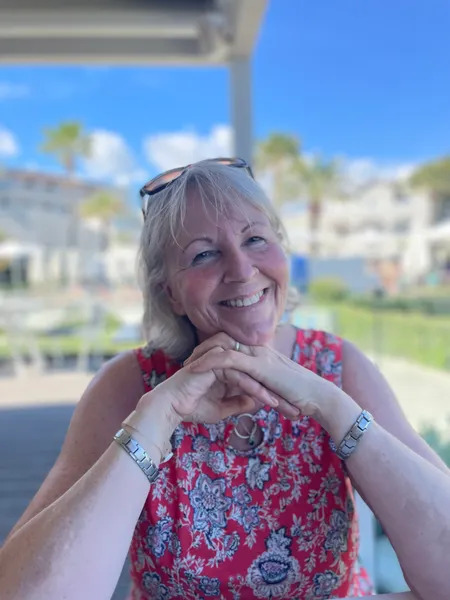 Smiling woman seated outdoors, wearing a red floral top, with sunglasses on her head.