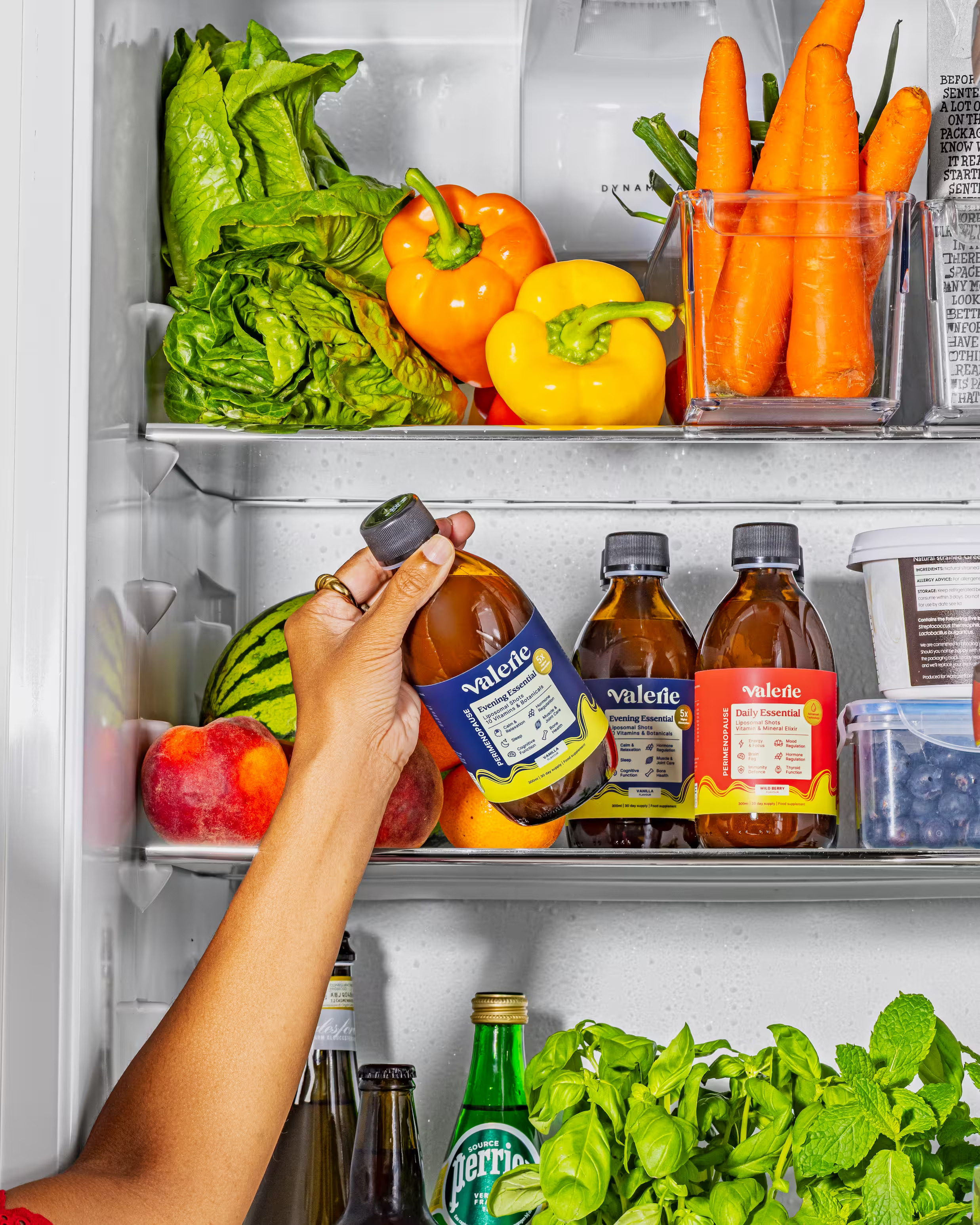 A hand reaching into a refrigerator full of vegetables, fruit, and bottled beverages.