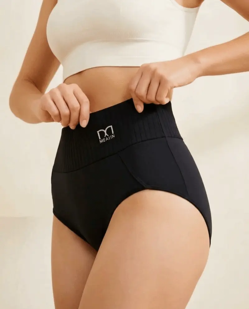 A person in a white top pulls up the ribbed waistband of black high-waisted underwear.