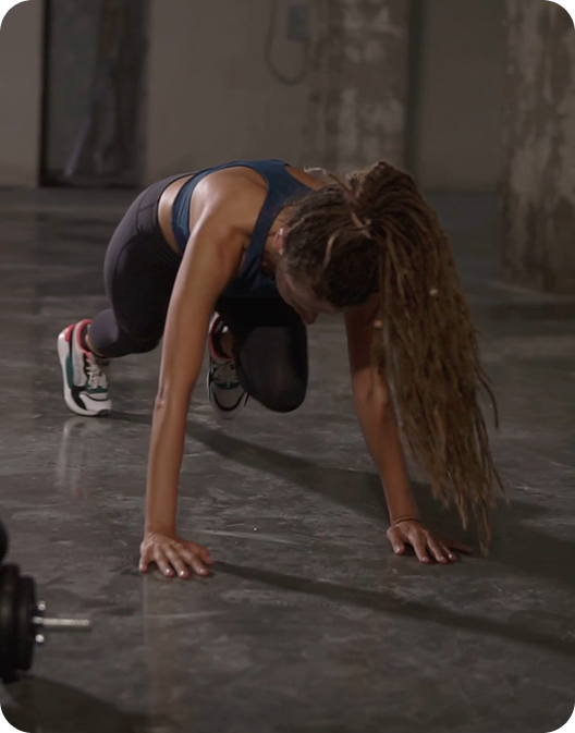 A woman with long dreadlocks in a plank position doing a mountain climber exercise on a concrete floor.