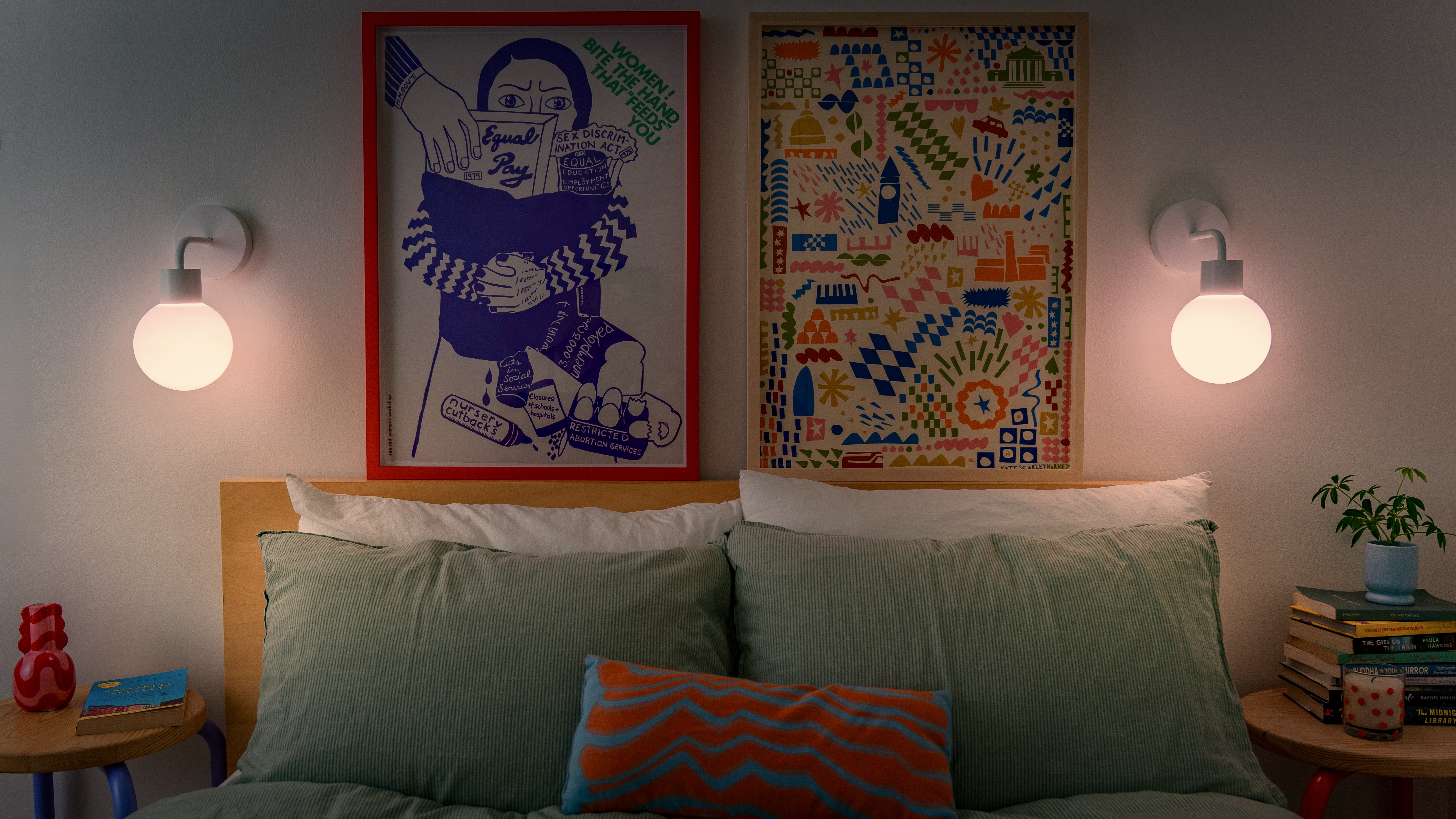 Bed with green pillows, two wall lamps, and colorful framed art above.