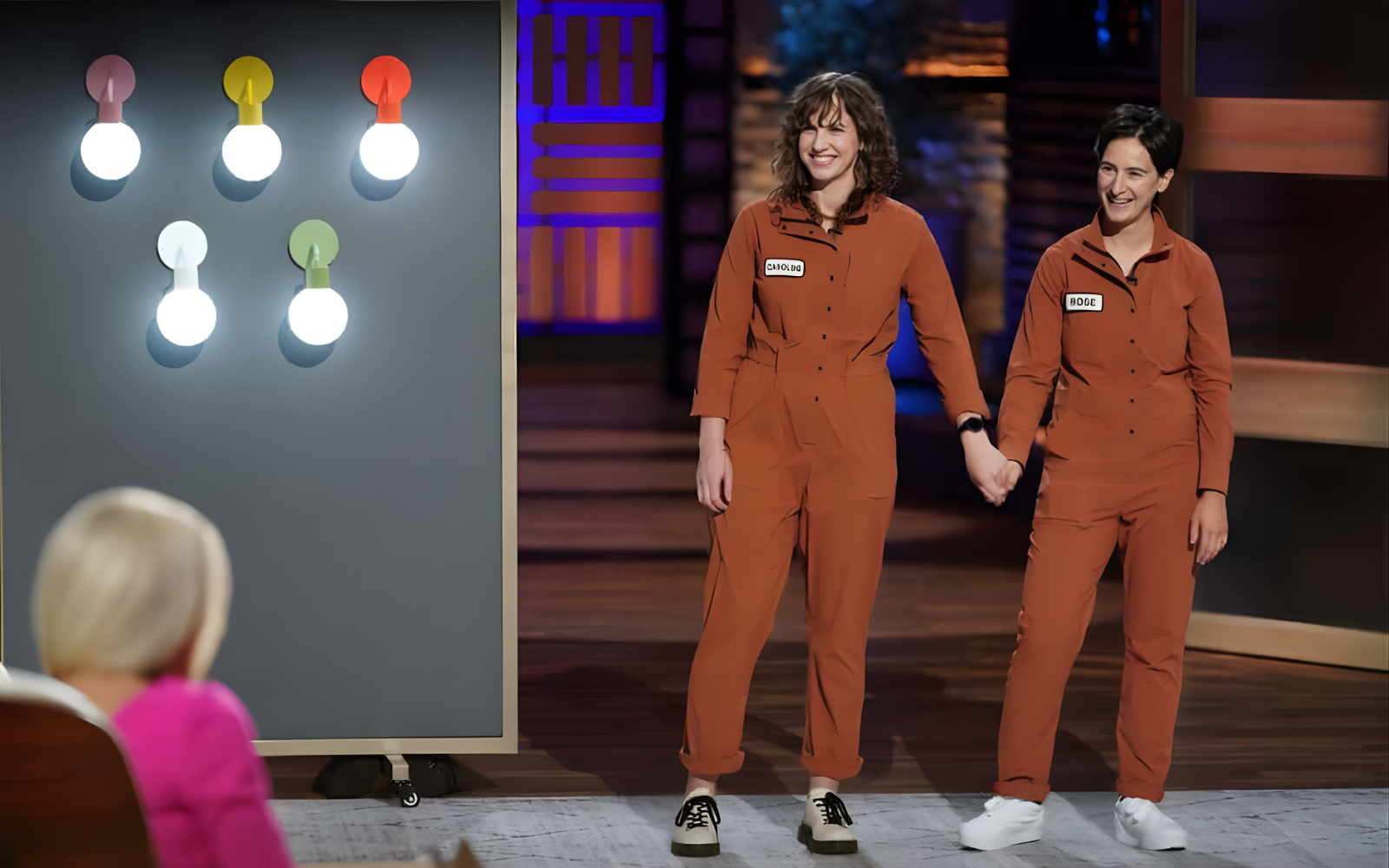 Two people holding hands in orange outfits, standing near a board with light bulbs.