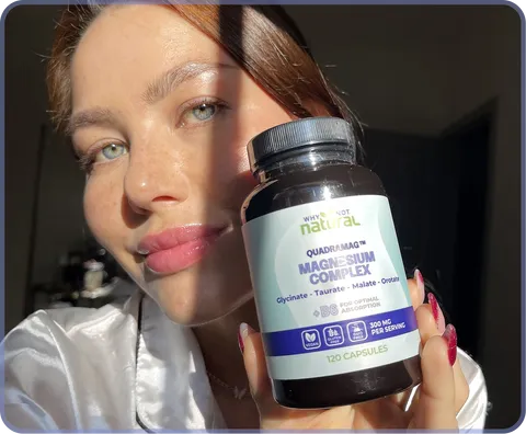 A woman with blue eyes holds a bottle of Why I'm Not Natural Magnesium Complex supplements.