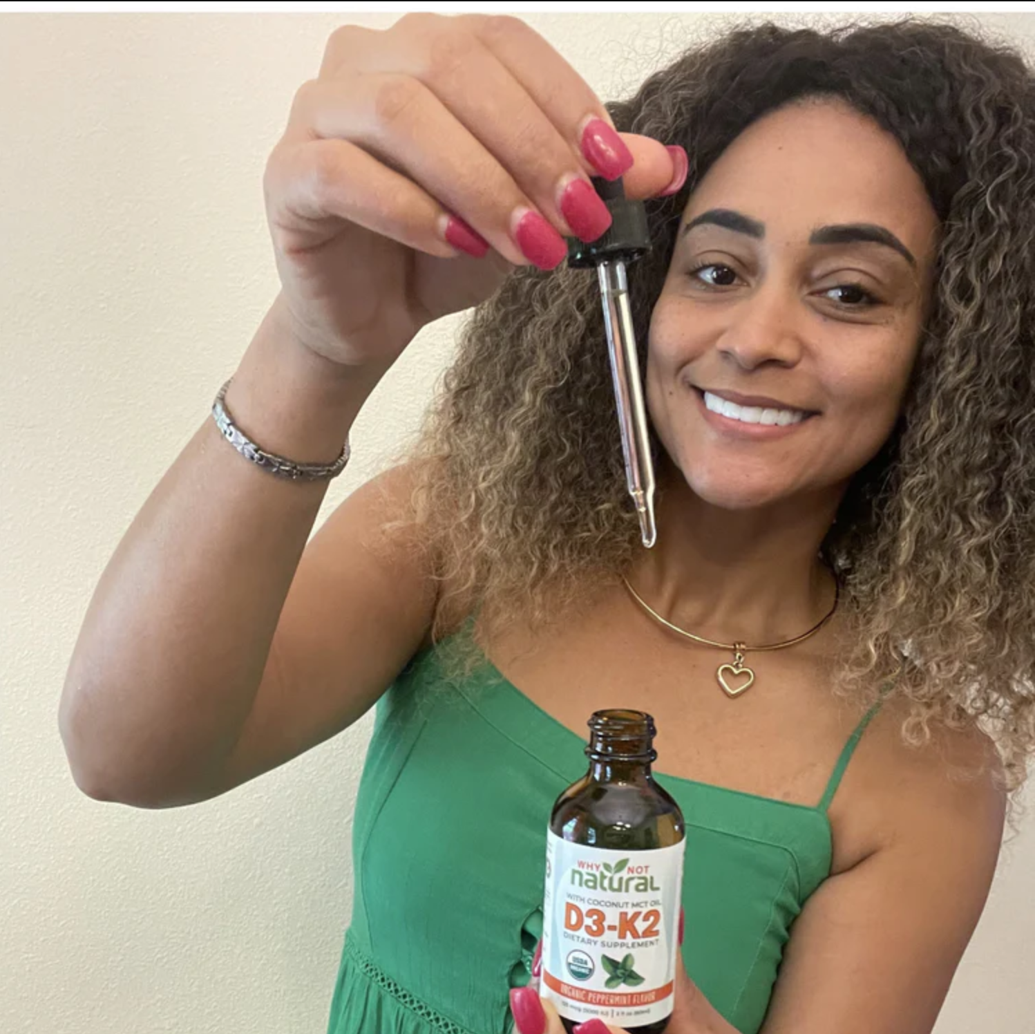 A smiling woman holds a bottle of Why Not Natural D3-K2 dietary supplement and a dropper.
