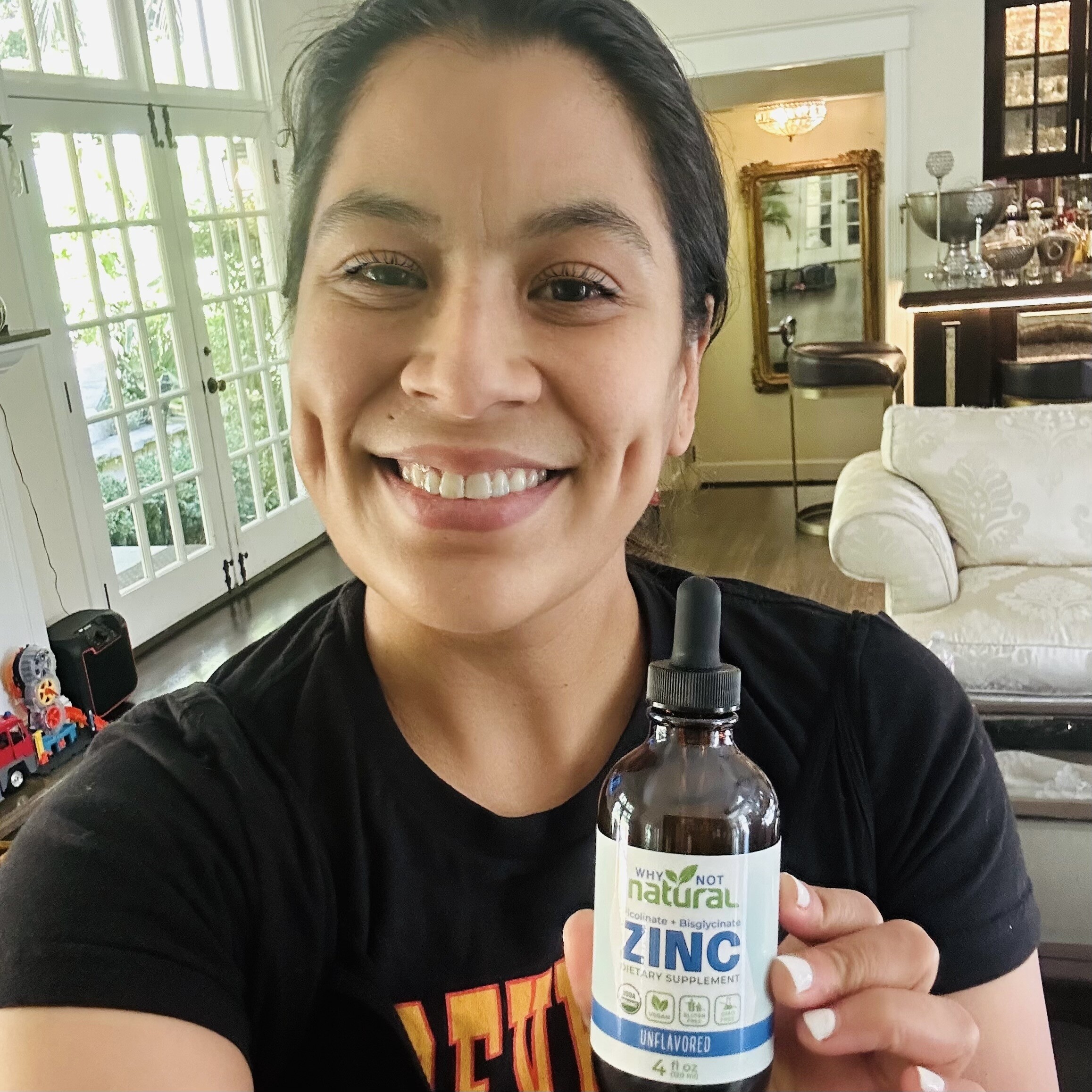 A smiling woman holds up a bottle of Why Not Natural liquid zinc supplement inside a home.
