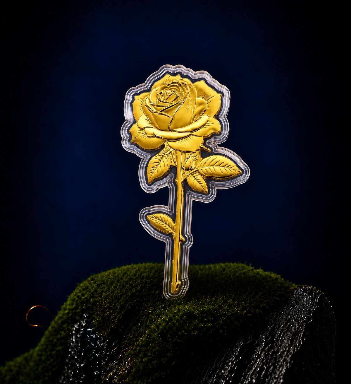 A gold, rose-shaped coin in a clear case, standing on a mossy surface against a dark background.
