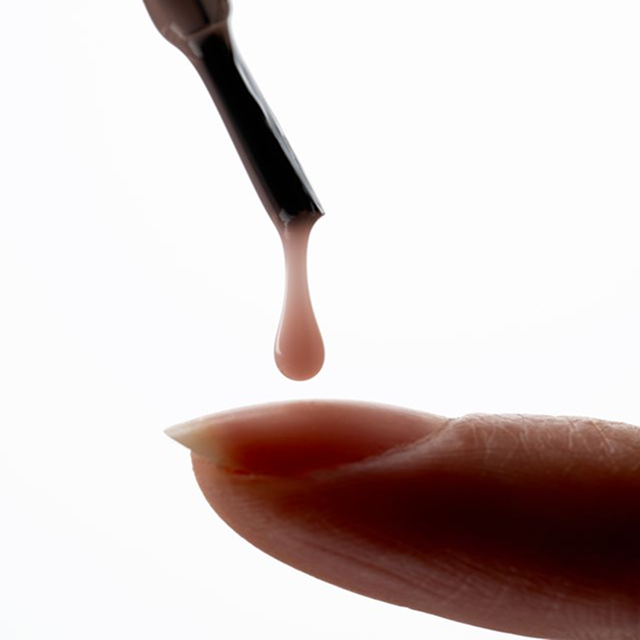 A close-up of a drop of nude nail polish falling from a brush onto a fingernail.